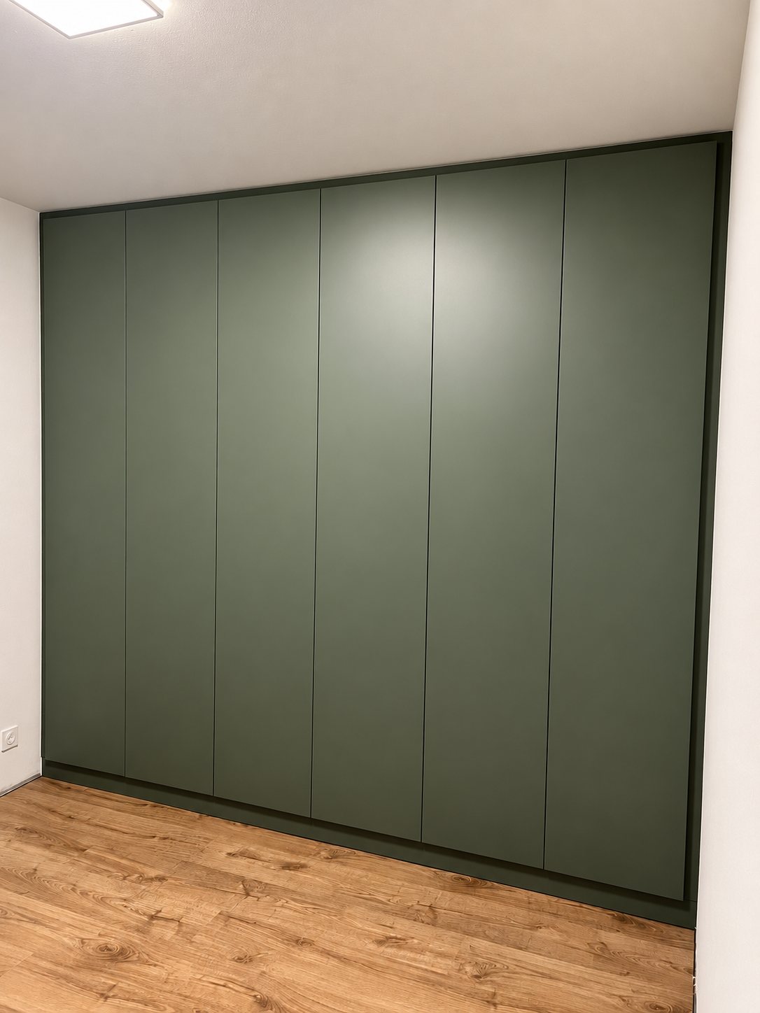 Un grand placard encastré vert kaki à six portes sans poignées, installé dans une pièce aux murs blancs avec un parquet en bois clair.