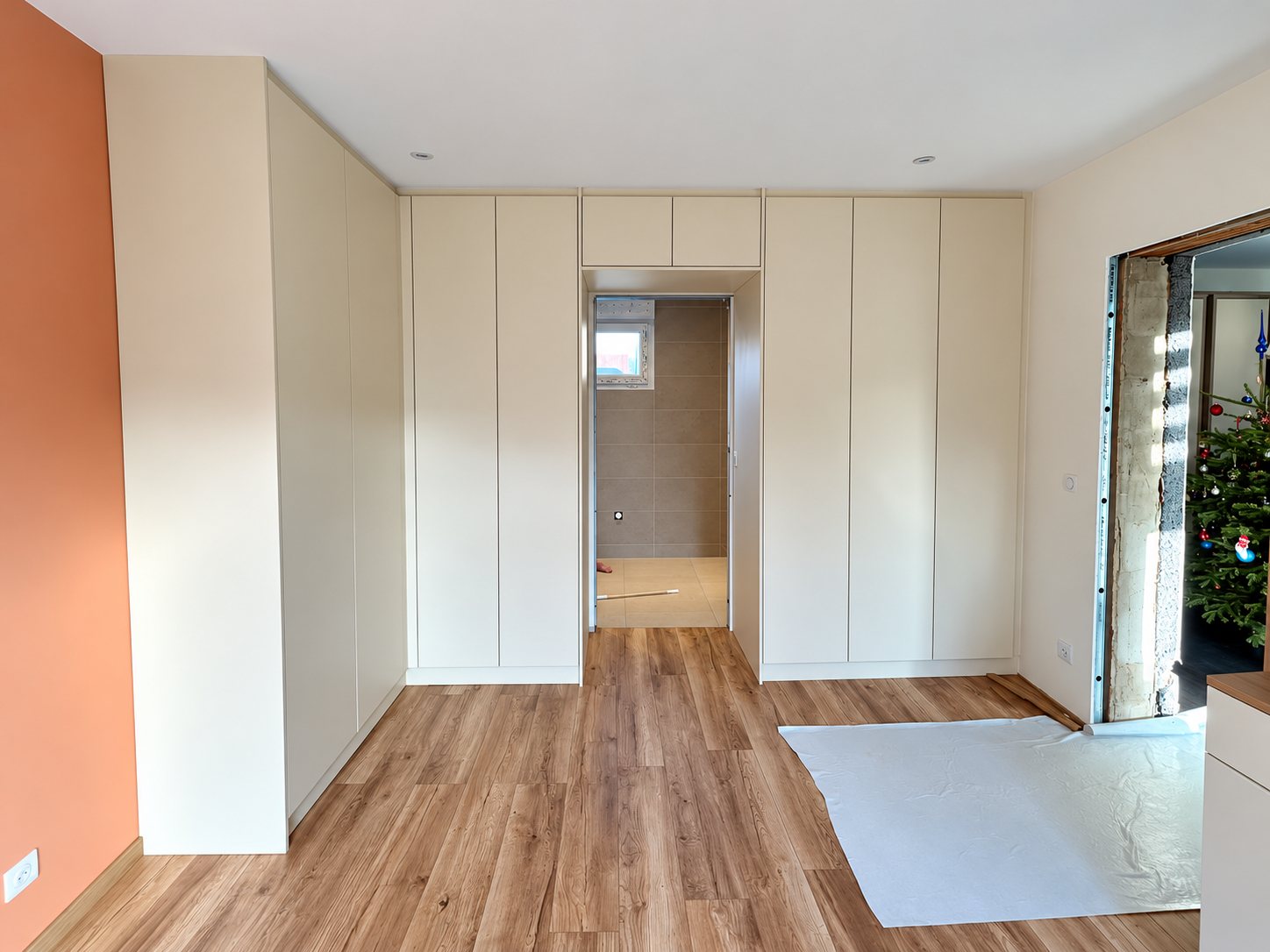 Une chambre rénovée avec des placards sur mesure beiges de part et d'autre d'une porte menant à une salle de bain carrelée, un parquet en bois et un sapin de Noël visible à droite.