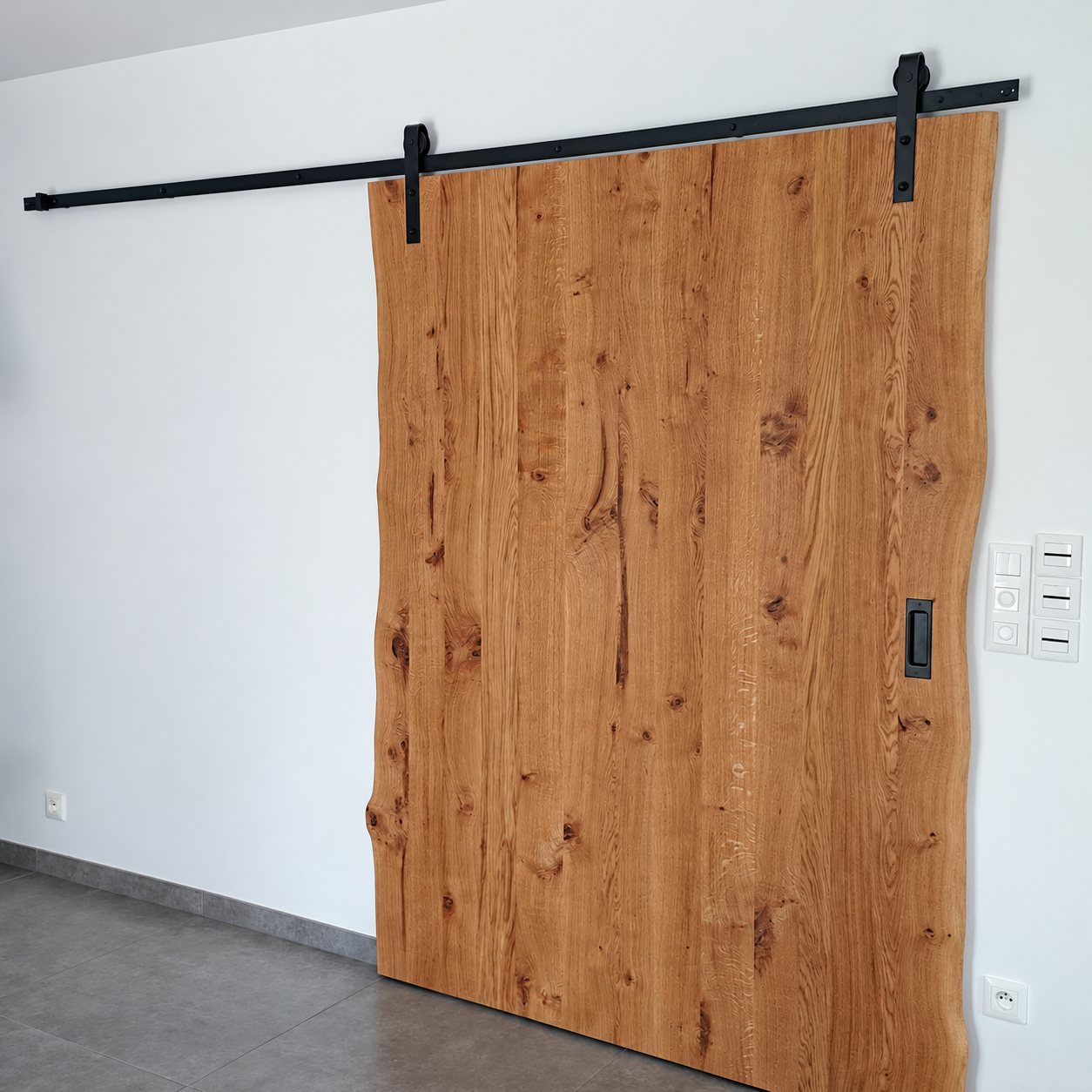 Une grande porte coulissante en chêne massif aux bords irréguliers est suspendue à un rail métallique noir, devant un mur blanc et un sol en carrelage gris.