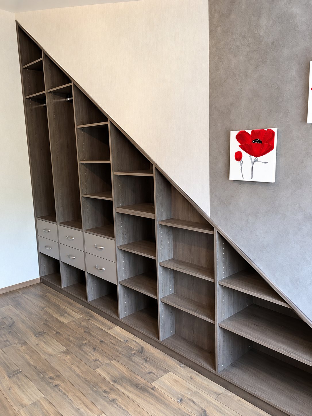 Meuble de rangement sur mesure installé sous un escalier, avec étagères, penderies et tiroirs en décor bois gris, près d'un tableau de coquelicot rouge.