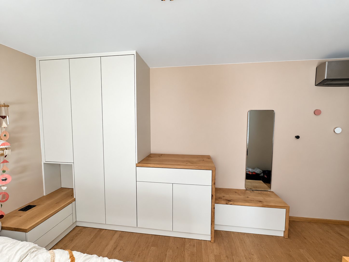 Dans une chambre aux murs roses, une armoire blanche sur mesure est associée à une commode et à des plateaux en chêne, avec un miroir étroit fixé au mur.