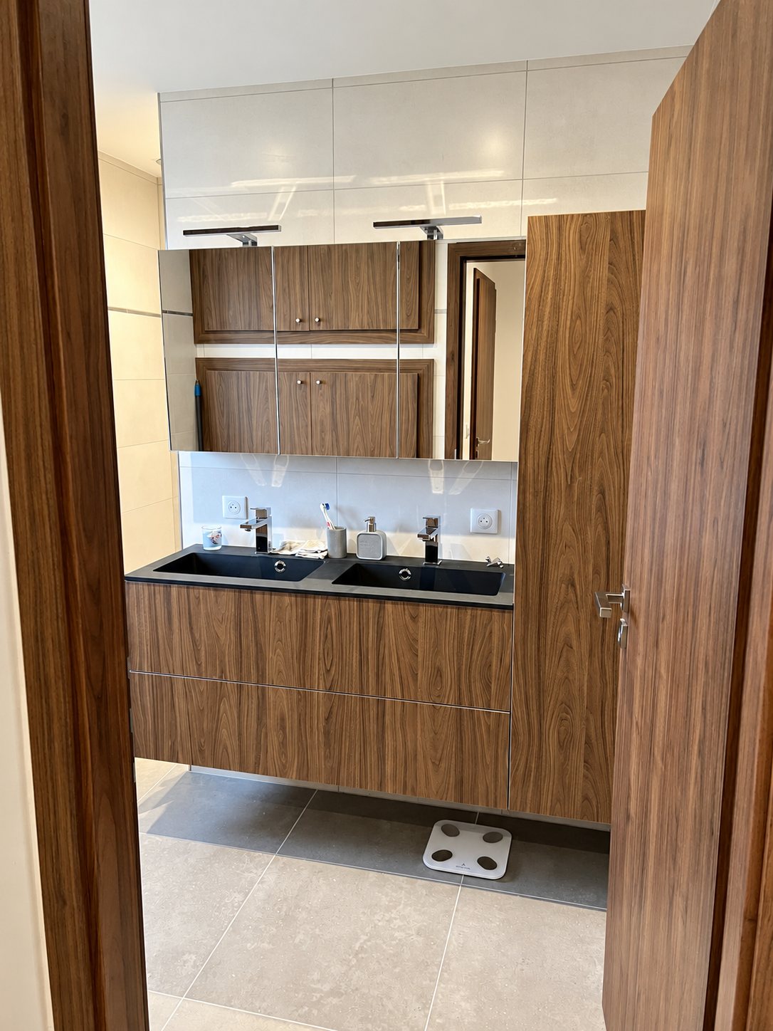 Salle de bain rénovée avec double vasque noire, meuble suspendu en noyer, armoire à miroir et grande colonne de rangement.