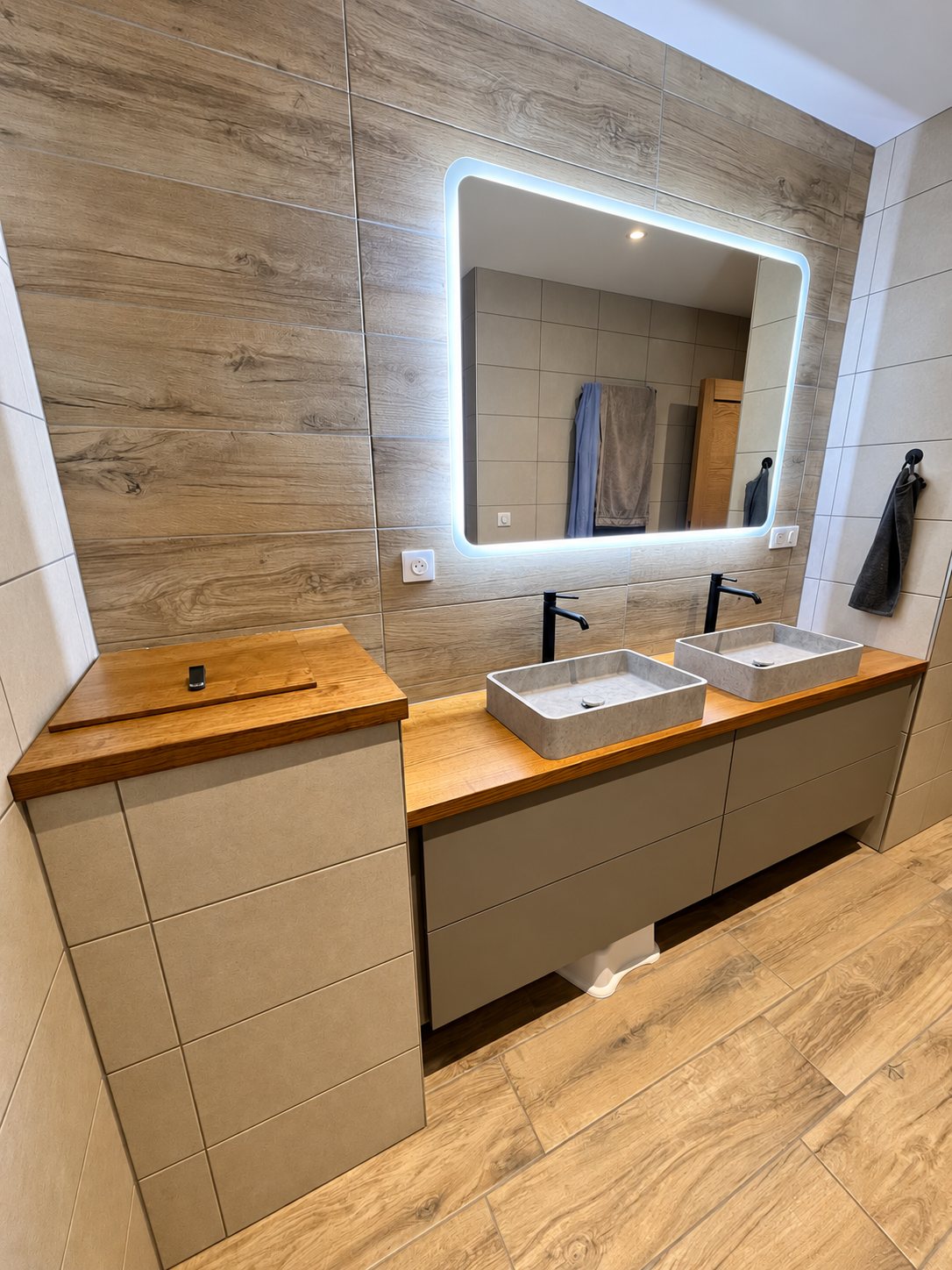 Salle de bain rénovée avec deux vasques rectangulaires grises posées sur un plan en bois, un miroir rétroéclairé et un carrelage aspect bois.
