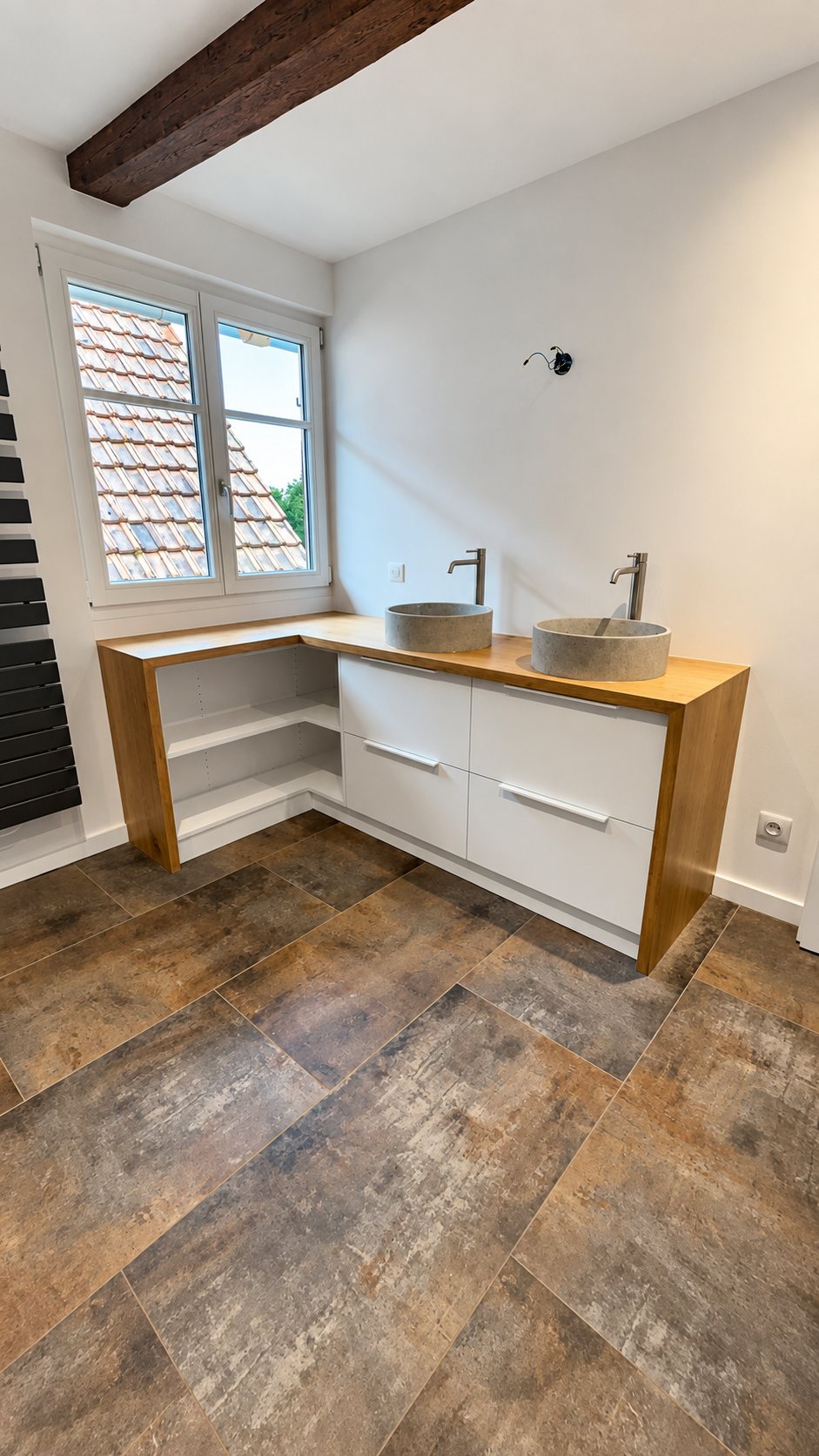 Salle de bains rénovée avec un meuble d'angle en bois et façades blanches, deux vasques en pierre à poser, un sol en grand carrelage et une fenêtre donnant sur un toit de tuiles.