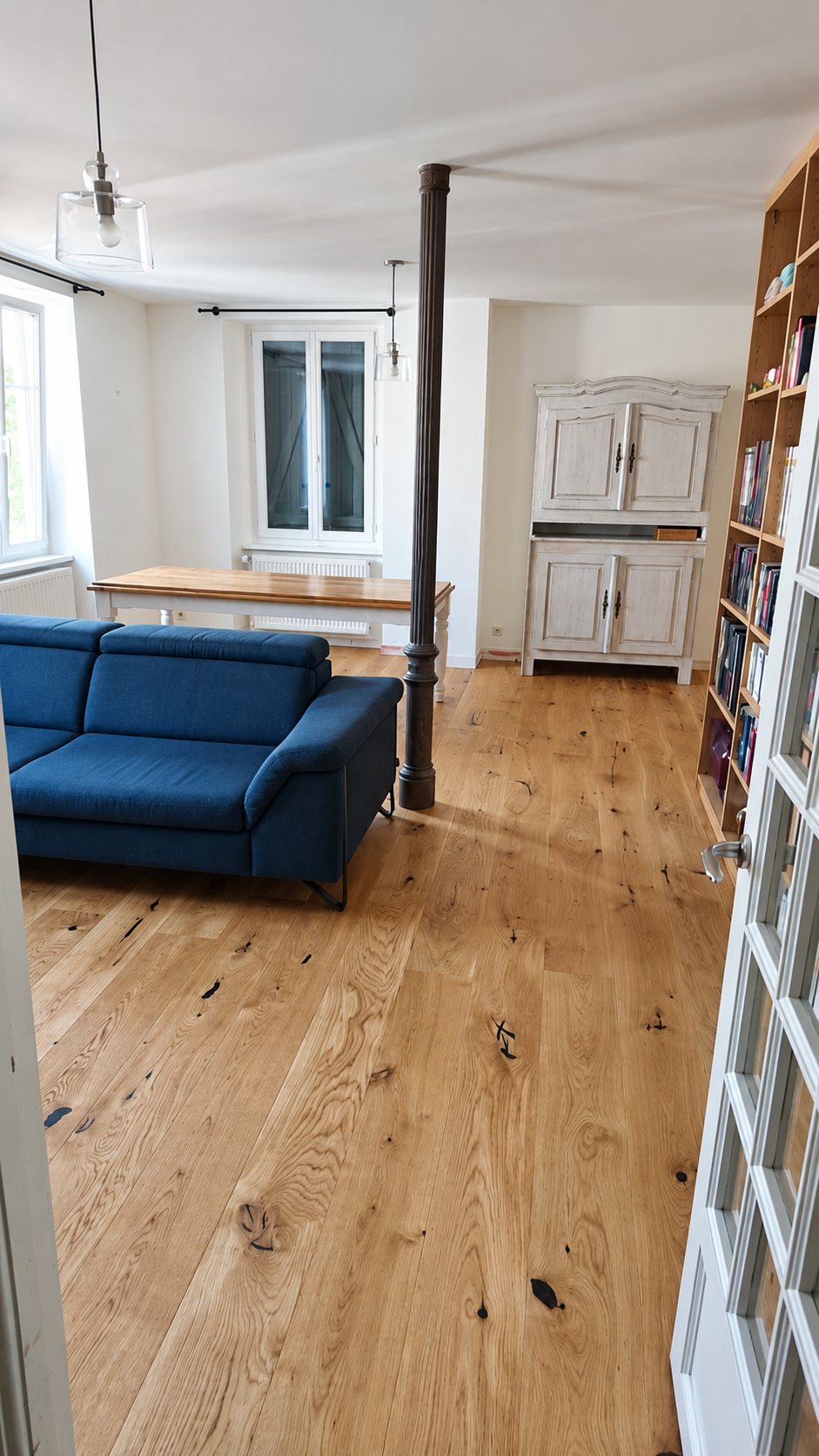 Salon lumineux avec un grand parquet en chêne, un canapé bleu, une colonne en fonte, une table en bois et une bibliothèque remplie de livres.