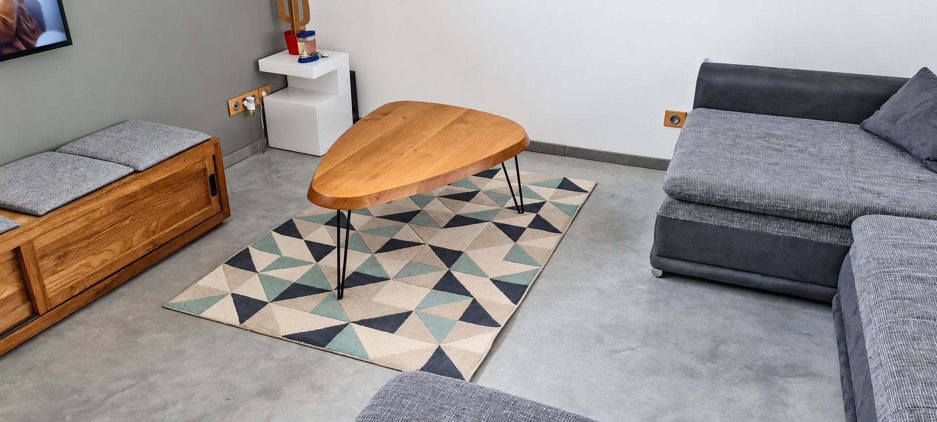 Une table basse en chêne de forme arrondie à pieds métalliques fins est posée sur un tapis à motifs triangulaires, entre un canapé gris et un meuble en bois, sur un sol en béton ciré.